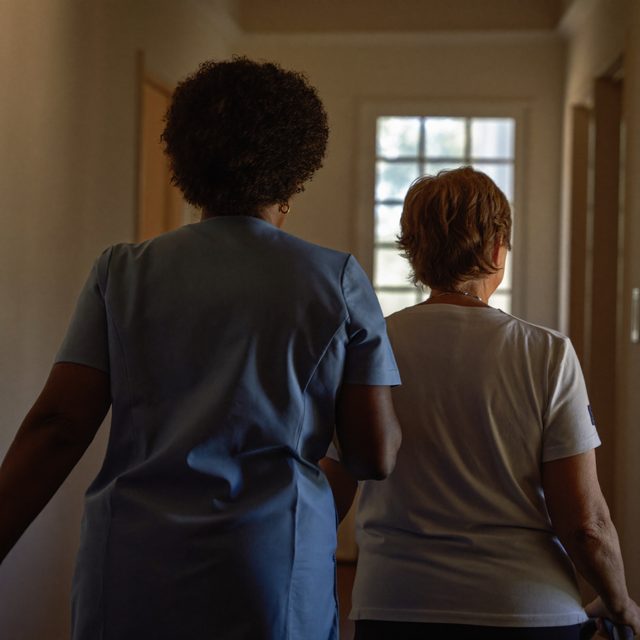 A caregiver walking beside an older woman along a sunlit hallway at home
